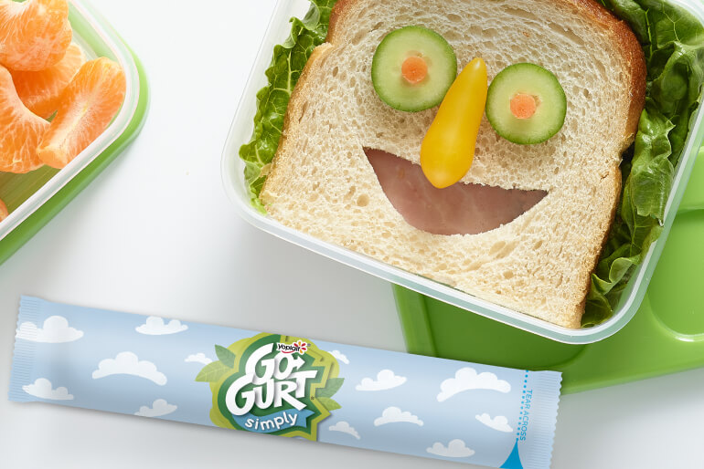 A Go-GURT tube sitting on a tabletop beside an open lunch box that contains a sandwich with ingredients arranged in a smiley face.