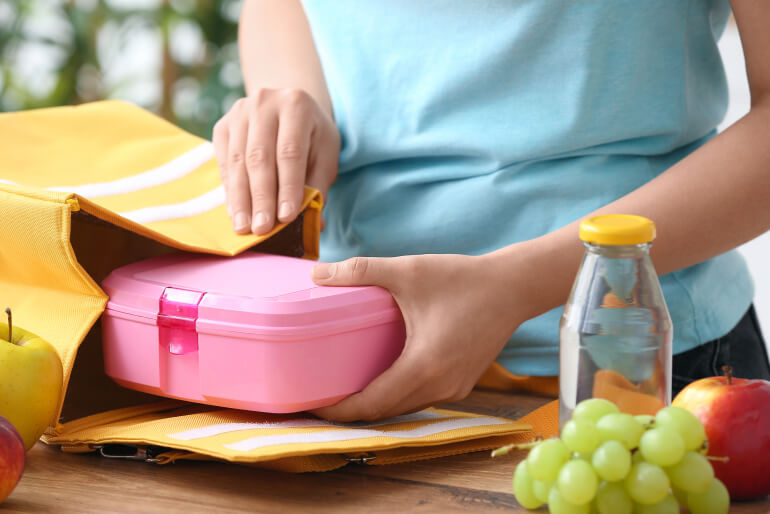 A woman packing a yellow bag with a pink lunch box with grapes and apples beside her.