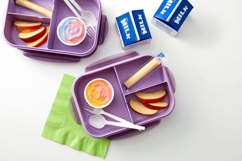 Two open purple lunch boxes with a pot of yogurt in each, apple slices and a sealed cheese string, beside two cartons of milk.