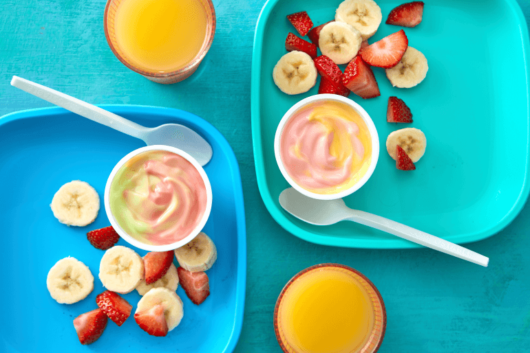 Two trays with a pot of yogurt on each, as well as strawberries and bananas, beside two glasses of orange juice.
